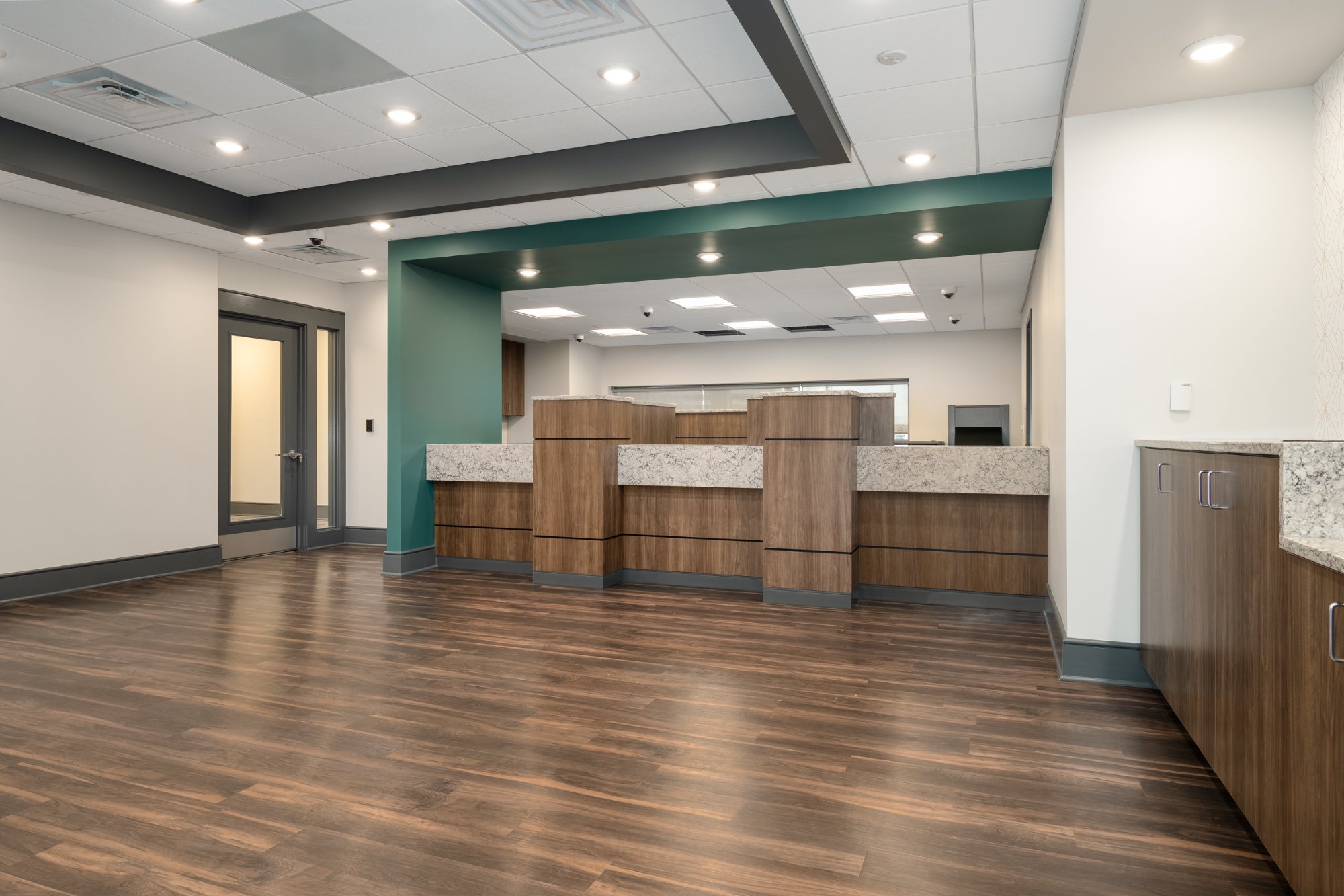 Bank lobby angled view of teller counter row with teal accent soffit dark hardwood floors and&nbsp;secured glass door
