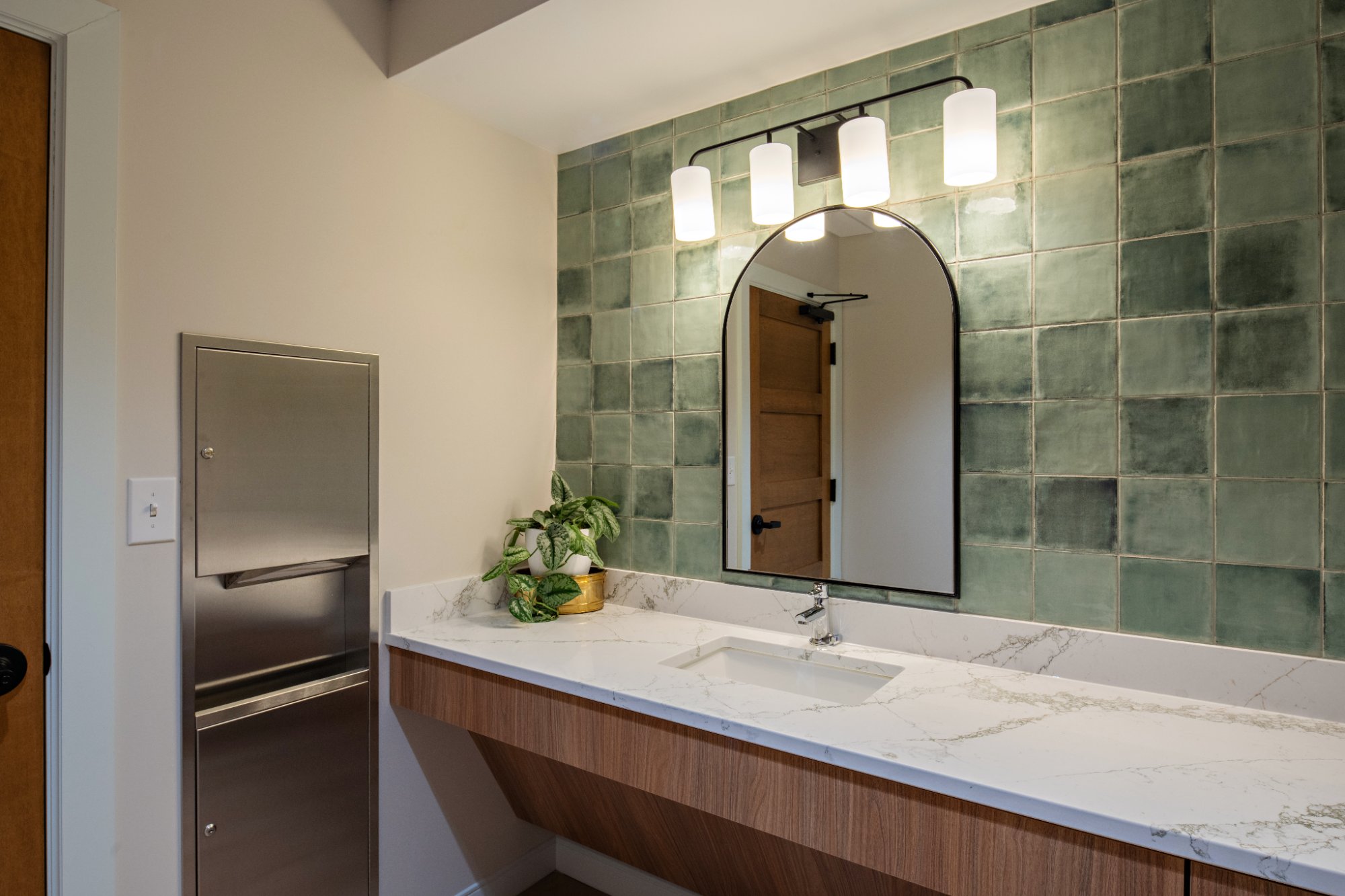 Dental-office-restroom-vanity-with-floating-walnut-cabinet-quartz-countertop-sage-green-tile-wall-arch-mirror-and-sconce
