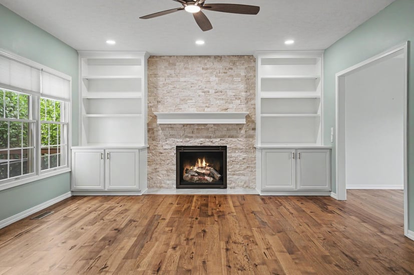 Living room with a stacked stone fireplace, white built-in shelving, and a ceiling fan after the remodel.