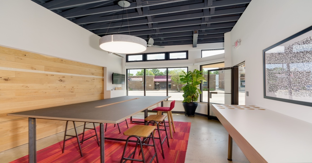 Collaborative meeting or community room with a large dark-topped worktable on sawhorse legs, mixed wood and red upholstered stools, a natural wood plank accent wall with a wall-mounted TV, and a drum pendant light overhead.